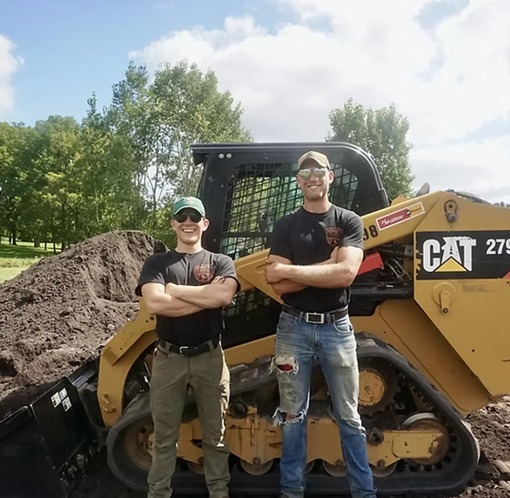 Two men wearing sunglasses and black shirts stand with arms crossed in front of a yellow CAT skid steer loader and a mound of dirt, with trees in the background. Kenneth Bros