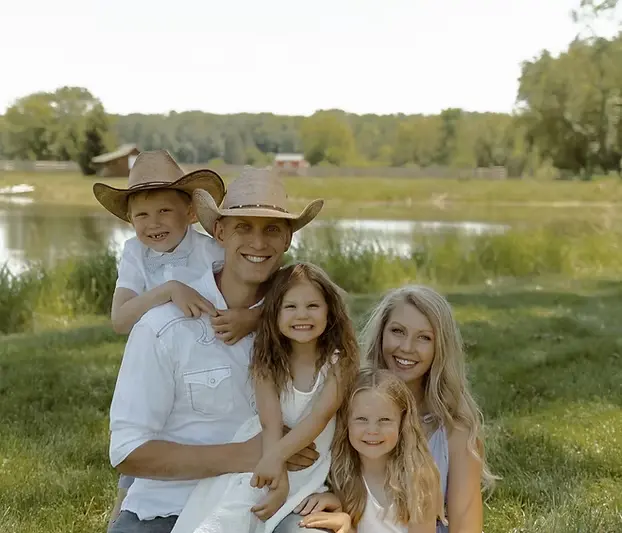A family of five, including two adults and three children, pose together outdoors by a pond, all smiling in casual clothes and cowboy hats—perfect for excavation contractors DeWitt MI enjoying a day off with loved ones. Kenneth Bros