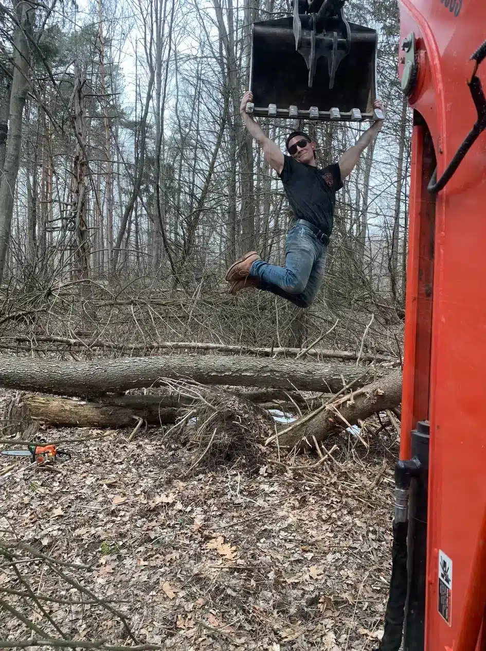 A man hangs with both hands from the bucket of an excavator in a wooded area, highlighting the demanding work often faced by a commercial demolition contractor in mid Michigan. Fallen trees and branches cover the ground. Kenneth Bros