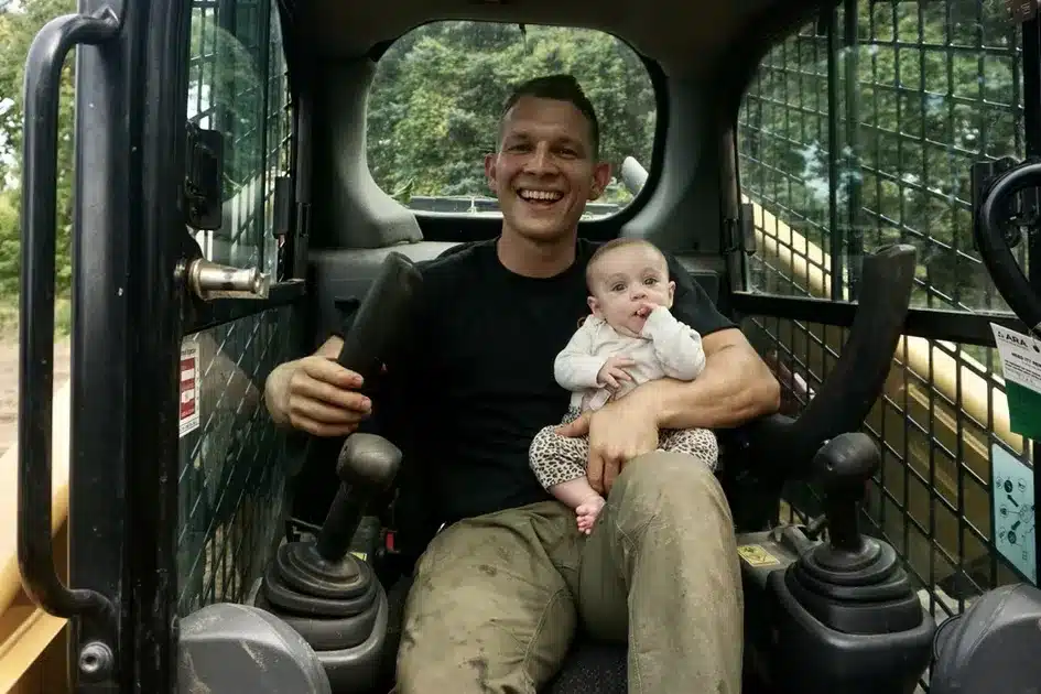 A man sits in the driver’s seat of a construction vehicle, smiling and holding a baby on his lap. Both are facing the camera—a charming moment from an excavation contractors DeWitt MI worksite, with trees visible through the window behind them. Kenneth Bros