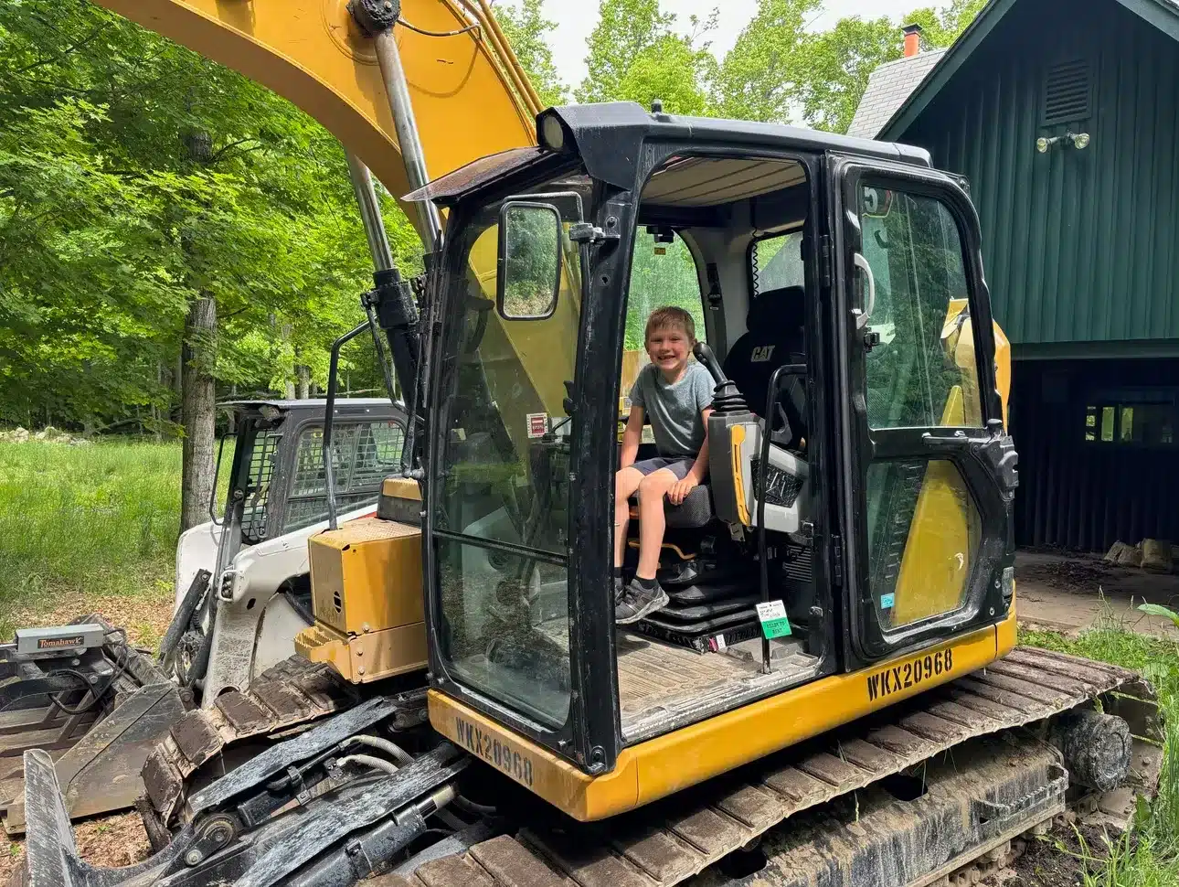 A young child sits in the operator’s seat of a yellow excavator, used for site preparation and grading services, parked outdoors near a green building and trees. Kenneth Bros