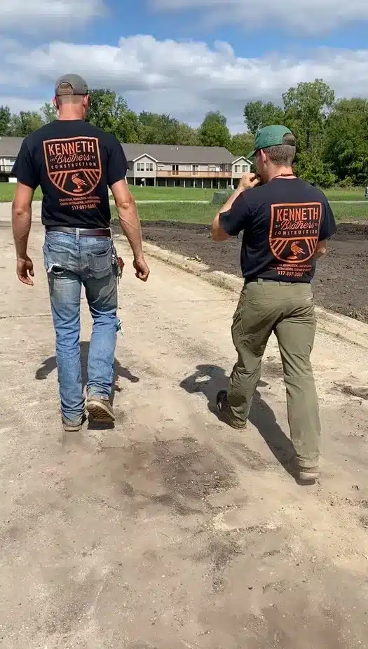 Two men in "Kenneth Construction" T-shirts walk on a dirt path near a grassy field and buildings, representing a trusted underground utility installation contractor MI. Kenneth Bros