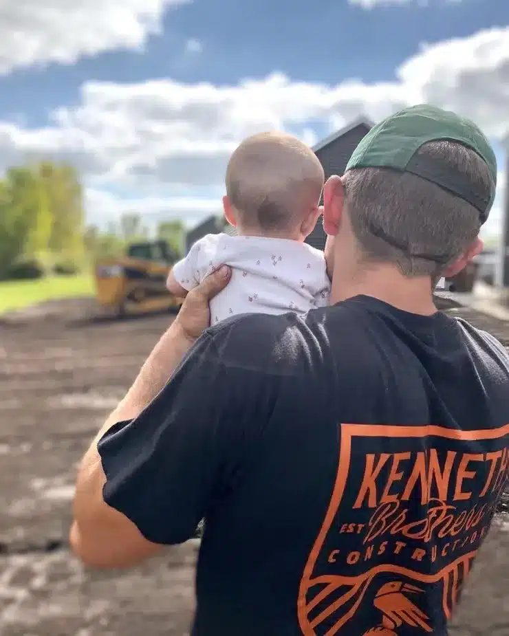 An adult in a green cap and black T-shirt holds a baby while standing outdoors near construction equipment on a sunny day, possibly at a site managed by an underground utility installation contractor MI. Kenneth Bros