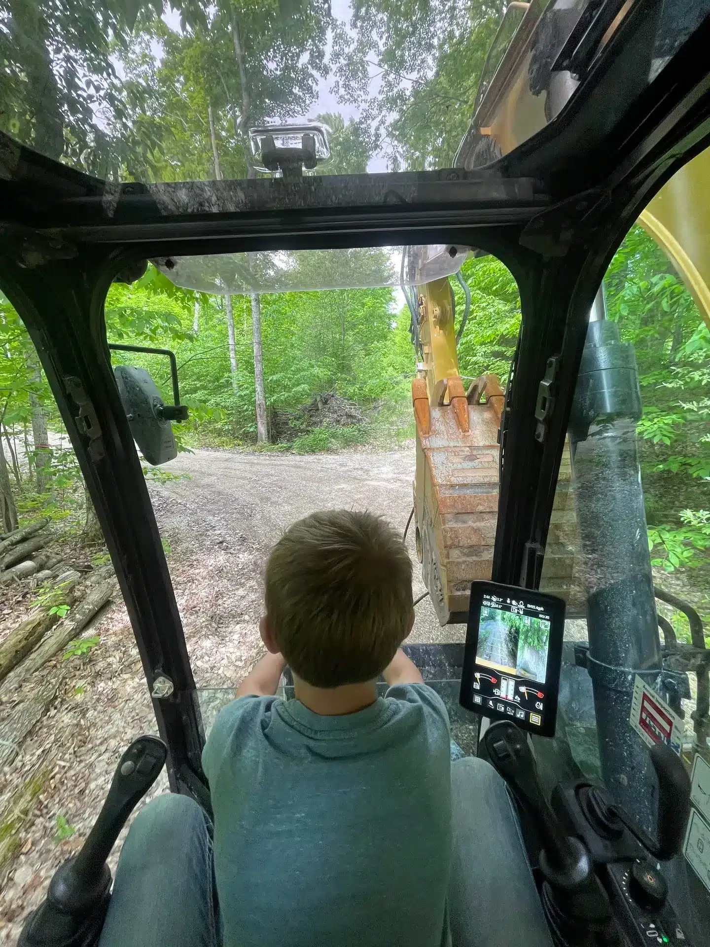 A child sits inside the cabin of an excavator, facing forward, with a forested path and control screen visible—perfectly capturing the spirit of site preparation and grading services in action. Kenneth Bros