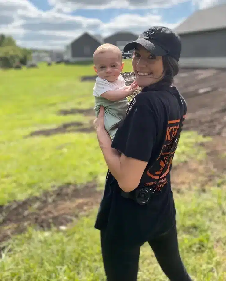 Woman in a black shirt and cap stands outside on grass, smiling and holding a baby who looks at the camera. Buildings—possibly future sites for commercial demolition contractor mid Michigan projects—and a cloudy sky are in the background. Kenneth Bros