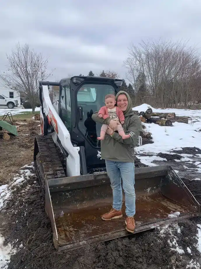 A person in a green jacket holds a baby and stands before a skid steer loader on muddy, snowy ground with bare trees—capturing the daily work of excavation contractors DeWitt MI in challenging weather. Kenneth Bros
