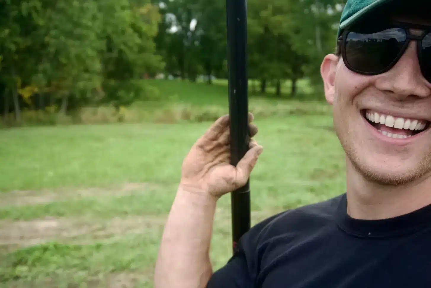 A person wearing sunglasses and a green cap smiles while holding a pole outdoors, with a grassy field and trees in the background—an ideal scene for excavation contractors DeWitt MI preparing for their next project. Kenneth Bros