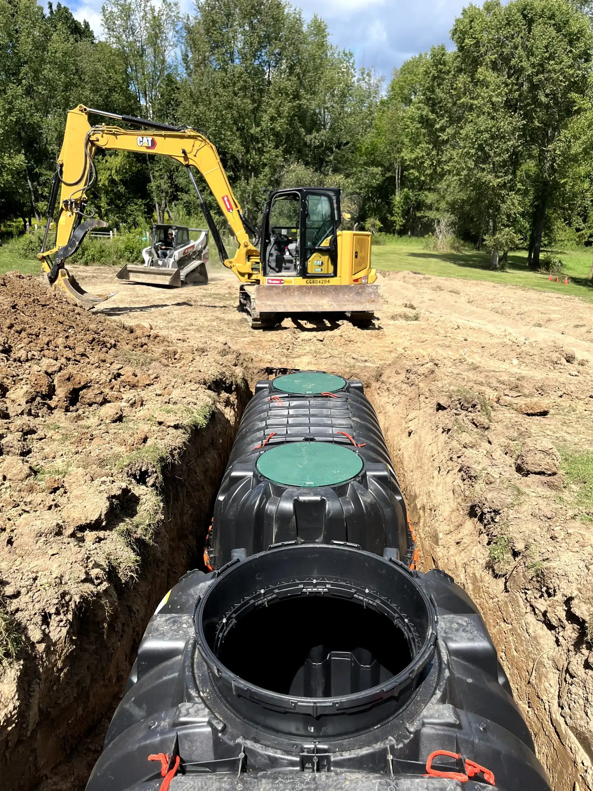 Excavator and skid steer near a trench with two large black plastic tanks being installed in a grassy area with trees in the background. Kenneth Bros