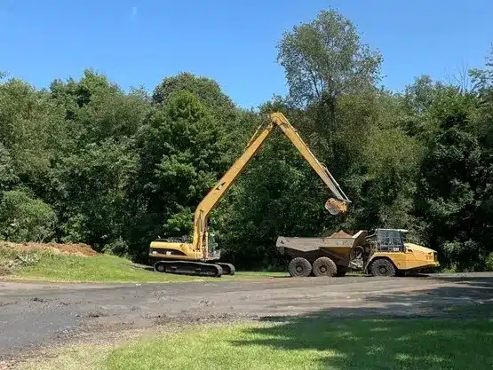 A yellow excavator with an extended arm loads dirt into a yellow dump truck on a paved area beside a wooded landscape. Kenneth Bros