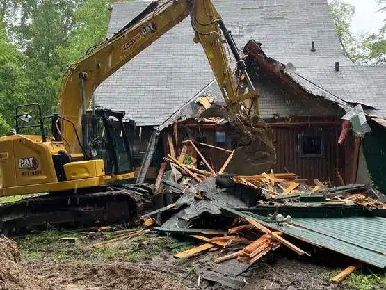 A yellow excavator demolishes part of a wooden house, with debris scattered on the ground in front of the machine. Kenneth Bros