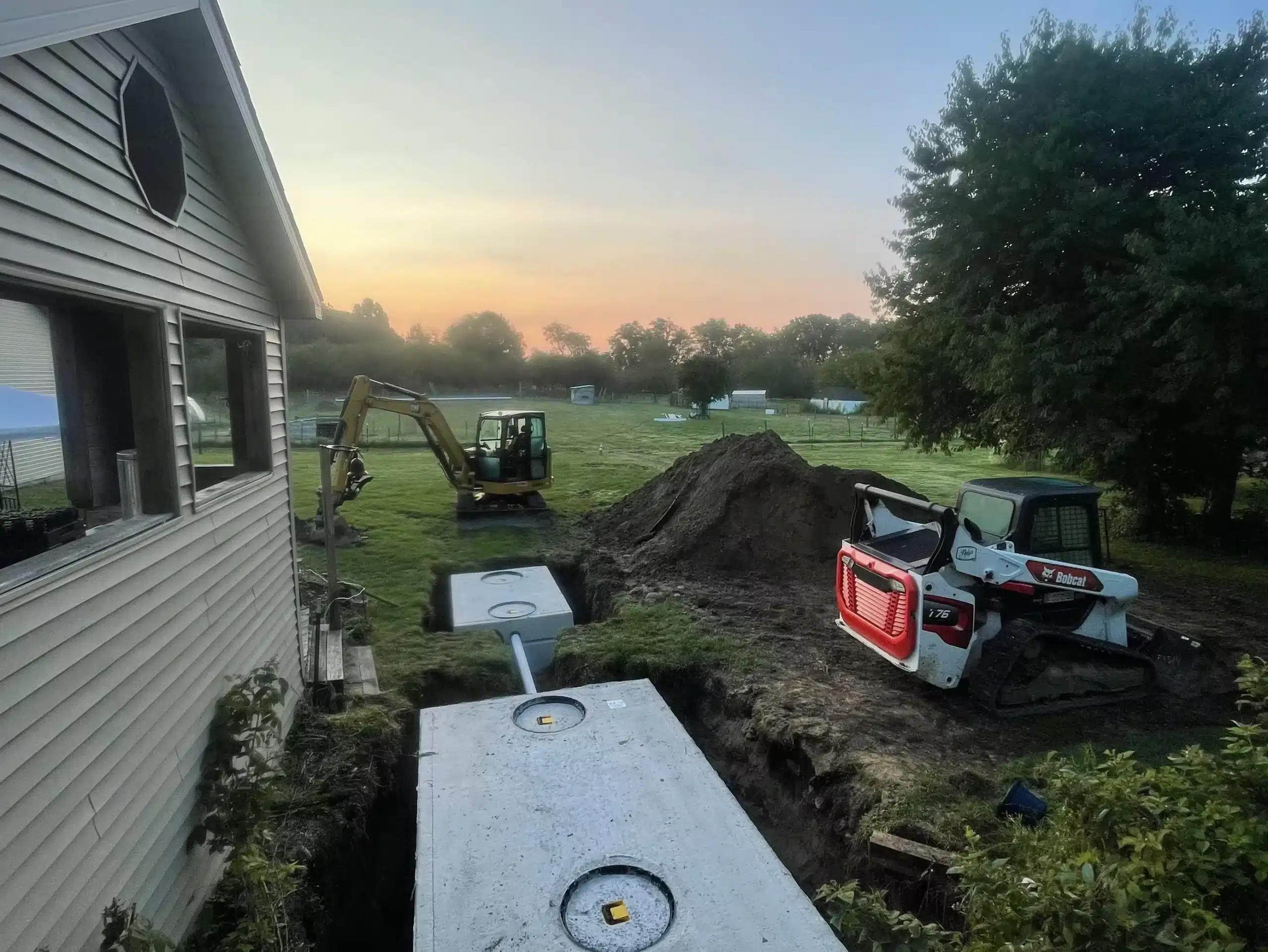 Construction equipment and workers install a concrete septic tank beside a house at sunrise, with a Bobcat loader and excavator on a grassy yard. Kenneth Bros