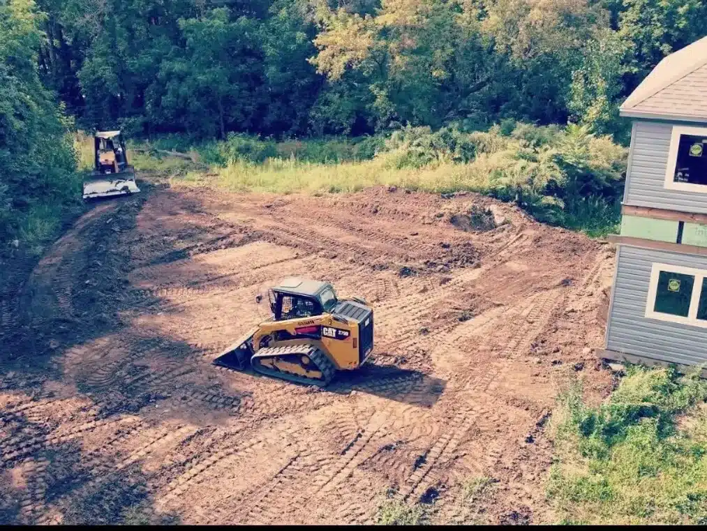 Two construction vehicles are leveling and clearing a dirt area next to a house, with trees and greenery in the background. Kenneth Bros