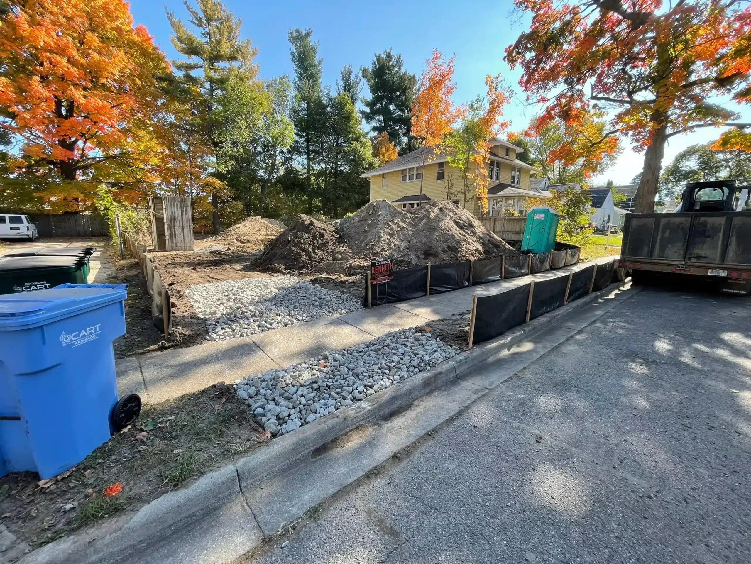Construction site with gravel and dirt piles, black silt fencing, a portable toilet, blue recycling bin, and a nearby house surrounded by autumn trees. Kenneth Bros