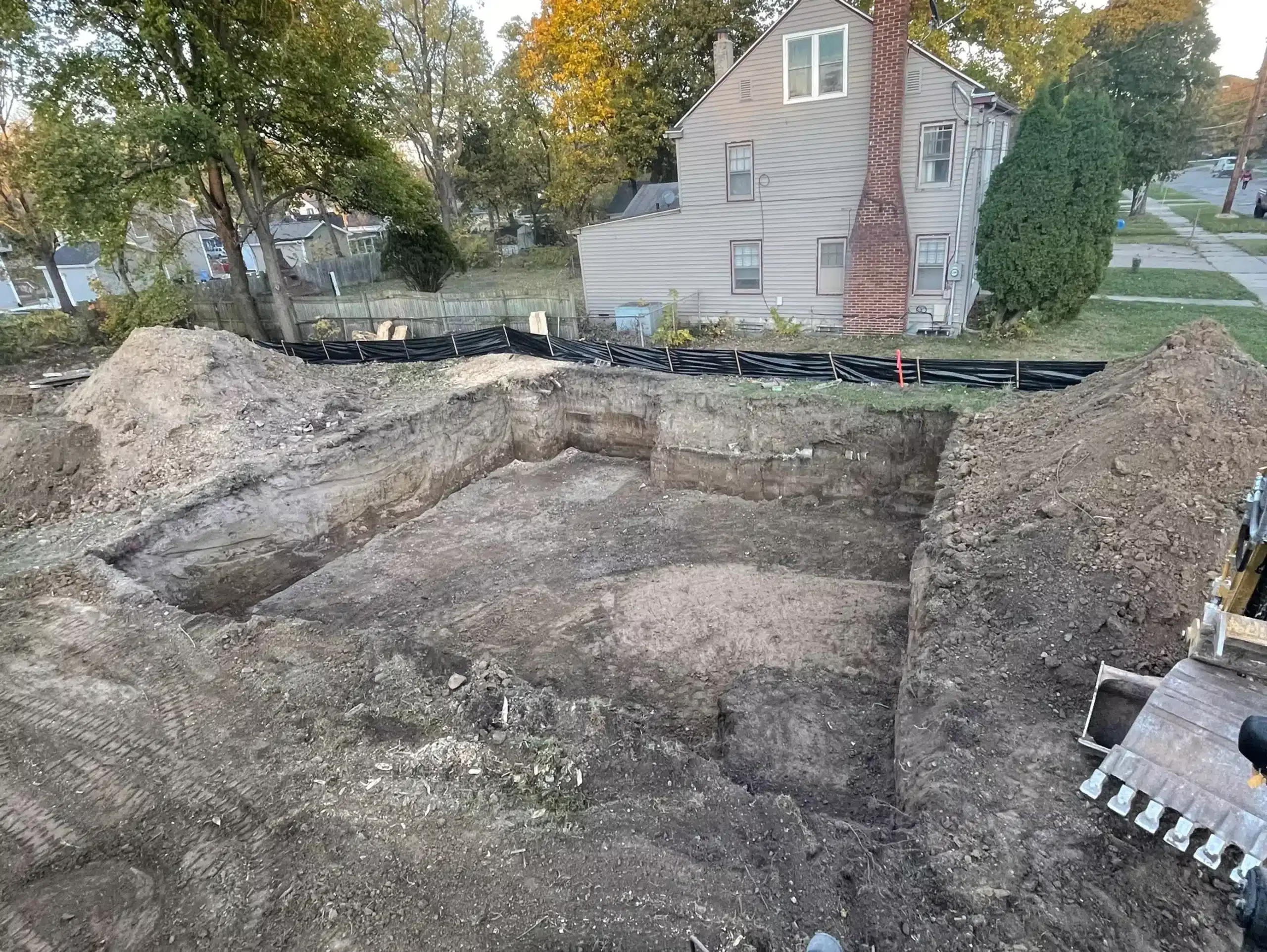 A large rectangular pit dug in the ground, likely for a building foundation, with a house and trees visible in the background. Kenneth Bros