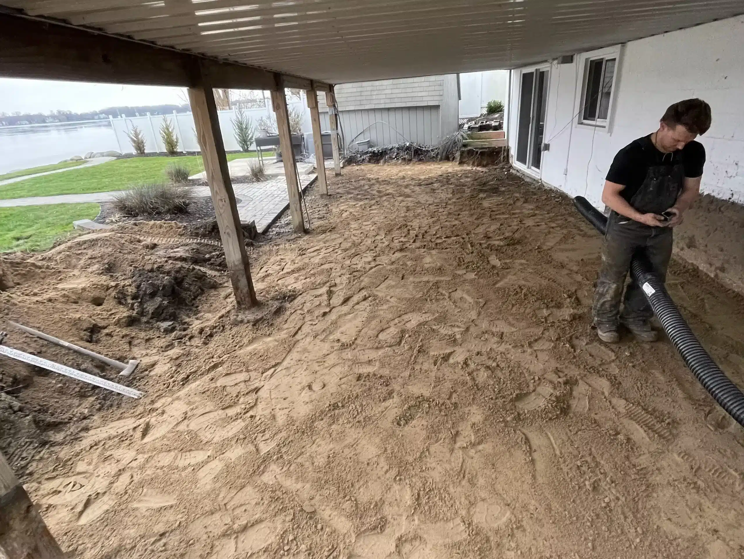 A person stands on a sandy surface under a patio roof, holding a large hose near a house; the area appears to be under renovation or landscaping. Kenneth Bros