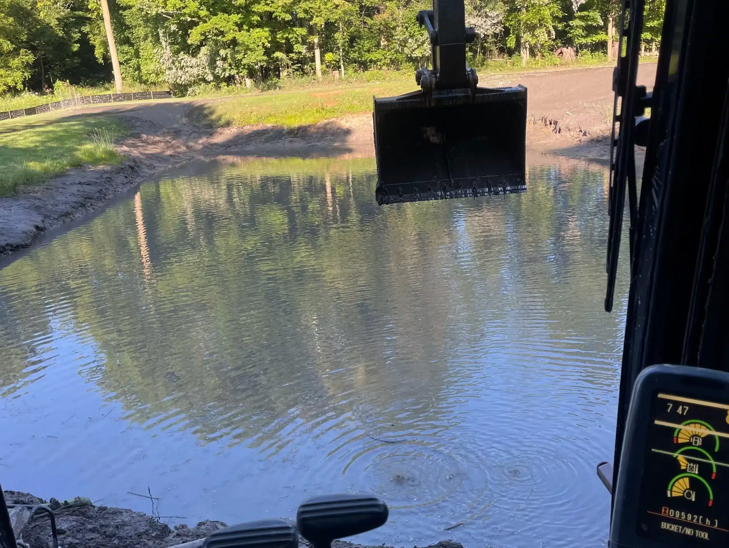 A construction excavator's bucket is suspended over a pond, with trees and grass visible in the background and a control panel in the lower right corner. Kenneth Bros
