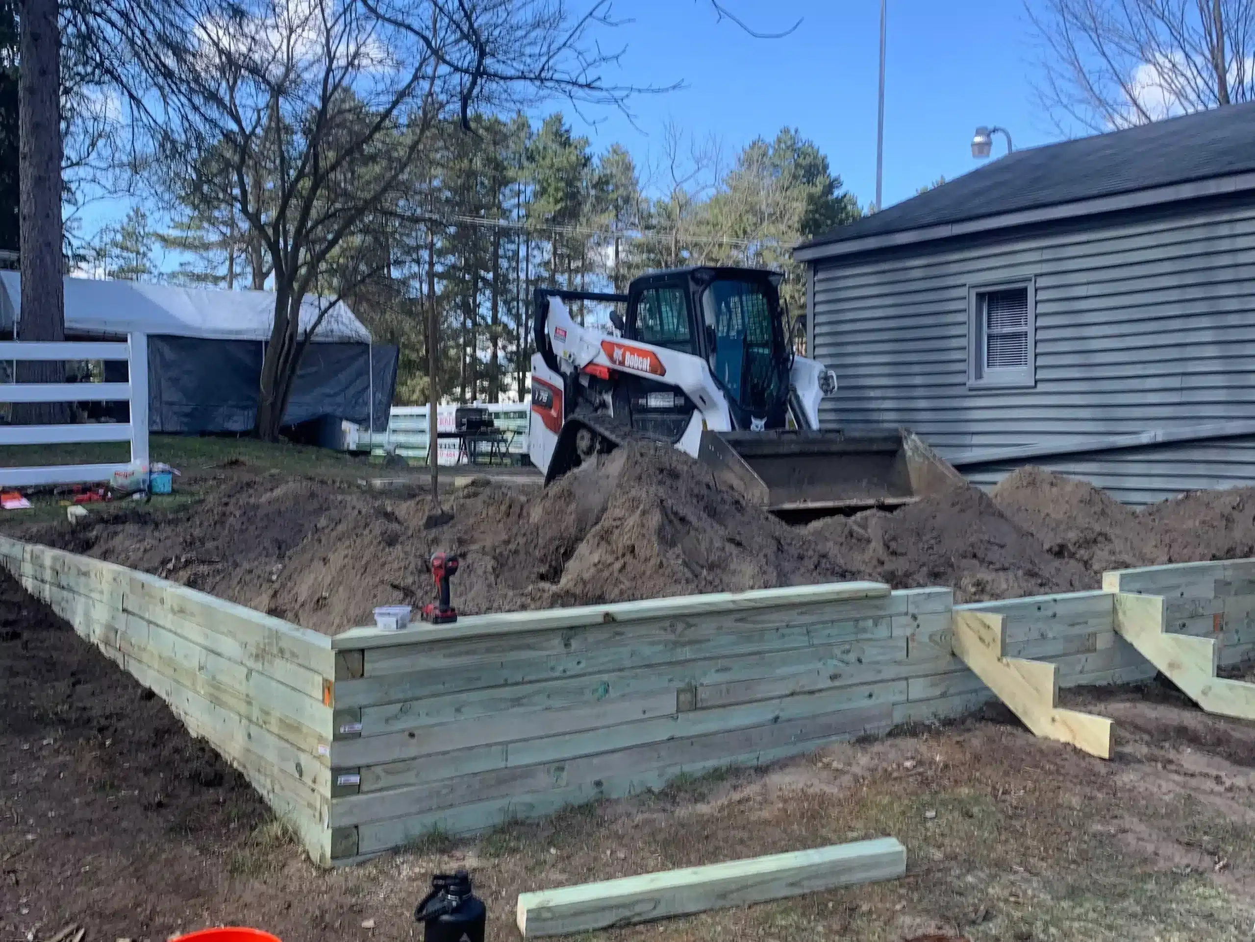 A skid steer moves dirt inside a partially built wooden frame next to a gray house; tools and building materials are visible around the construction site. Kenneth Bros