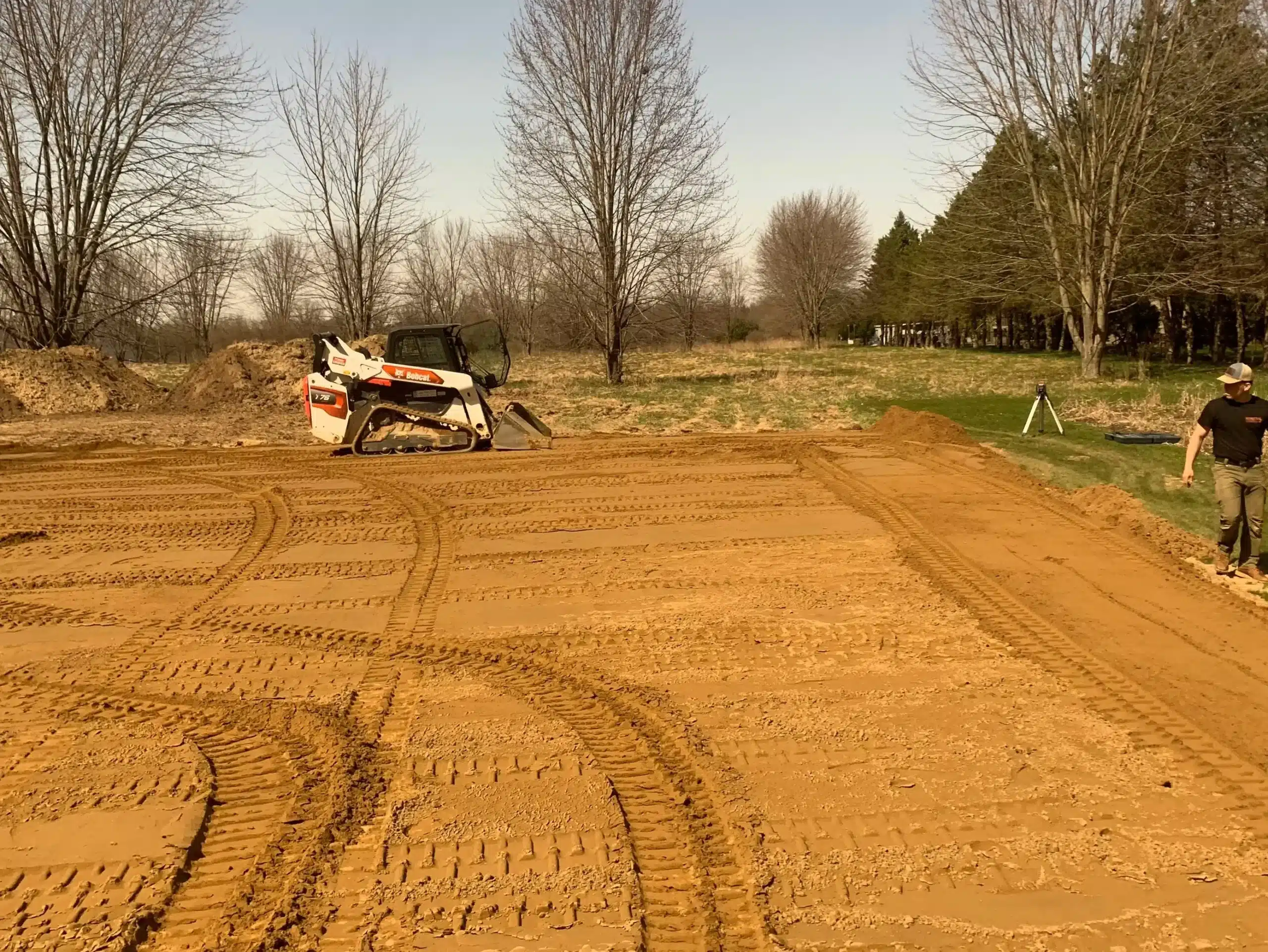 A construction site with leveled dirt, visible tire tracks, a compact track loader, and a person standing on the right side near trees. Kenneth Bros