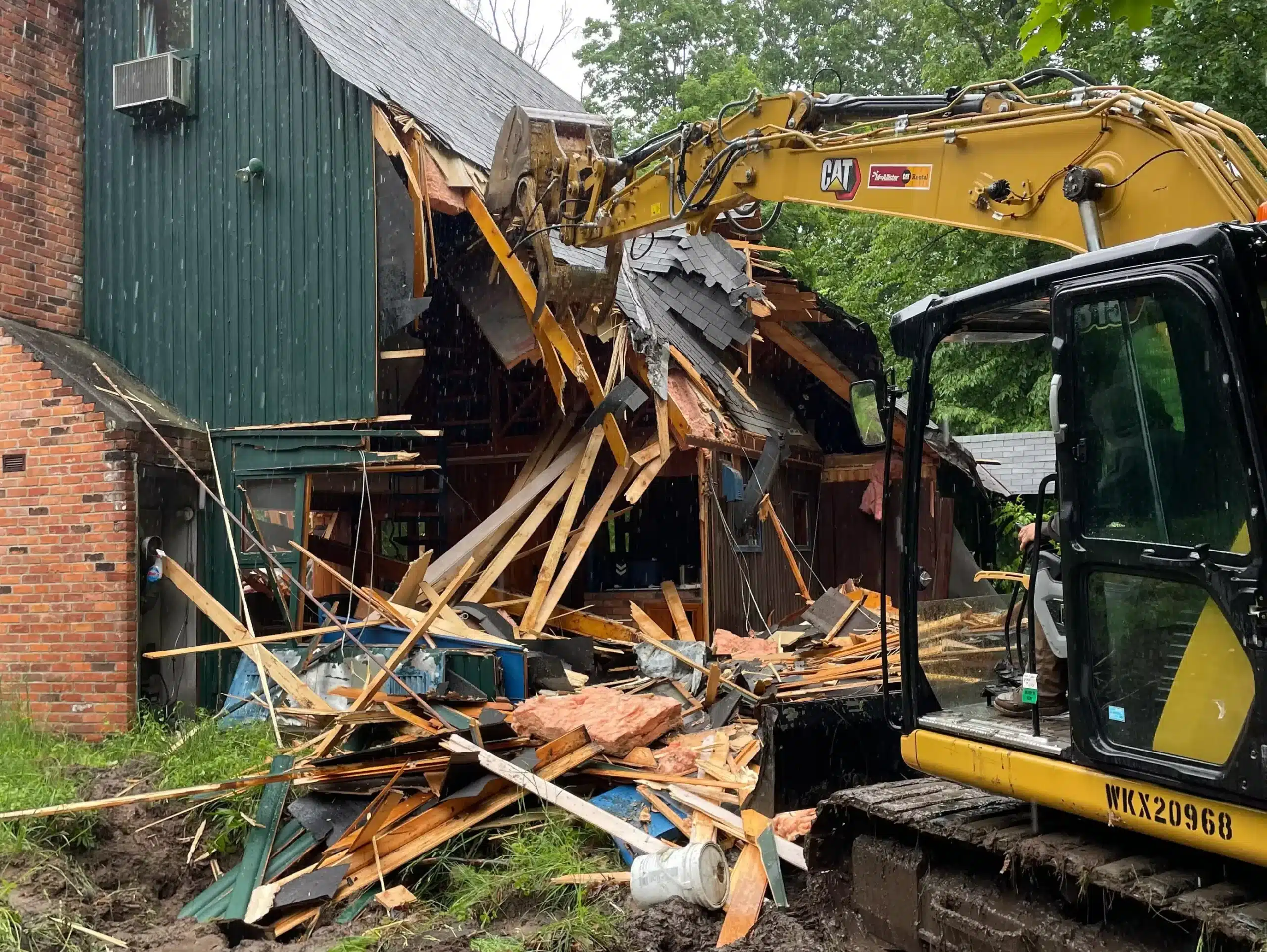 A yellow excavator is demolishing part of a wooden building, causing debris and wooden beams to collapse to the ground. Kenneth Bros