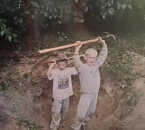 Two young boys stand in a dirt hole outdoors, each holding up a gardening tool above their heads; the scene resembles a site preparation and grading services project, with trees and grass visible in the background. Kenneth Bros
